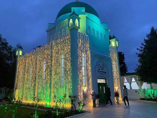 The Fazl Mosque, a white building with a green dome, covered in strings of gold lights and lit green at night